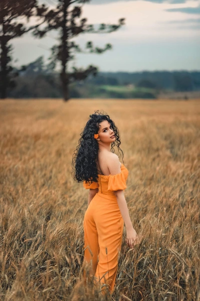a young woman with long black curly hair staying in the middel of the field wearing an orange jumpsuit
