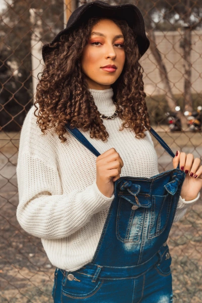 a young woman with black curly har wearing a whit knit pullover and a denim overall