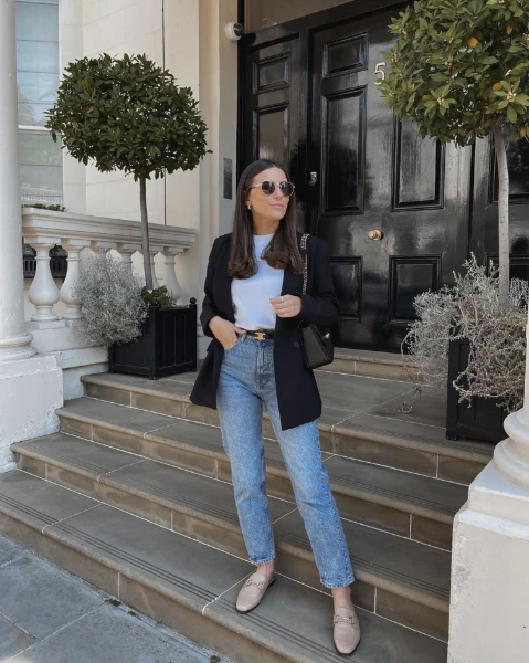a young woman staying on stairs in front of a house. she wears blue jeans, a white top, a black jacket and sunglasses