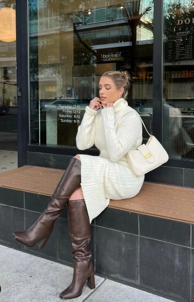 a blonde Woman sitting in front of a store with a white sweater dress and brown riding shoes